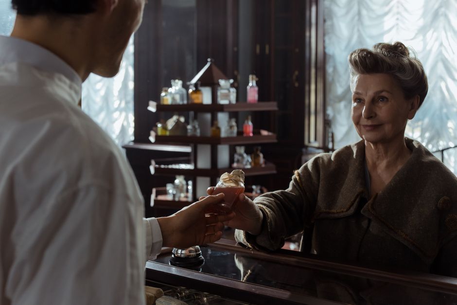 A senior woman receives a perfume bottle from a pharmacist inside a vintage pharmacy.
